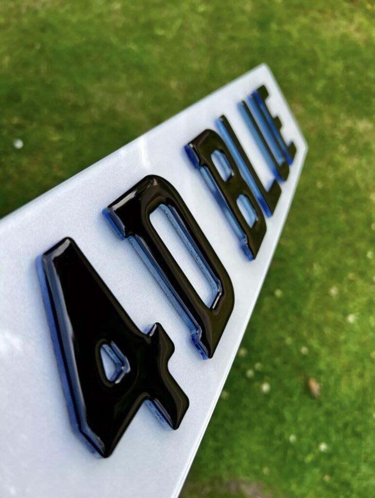 Close-up of 4D number plate letters with glossy black finish and blue gel backing on a white plate.